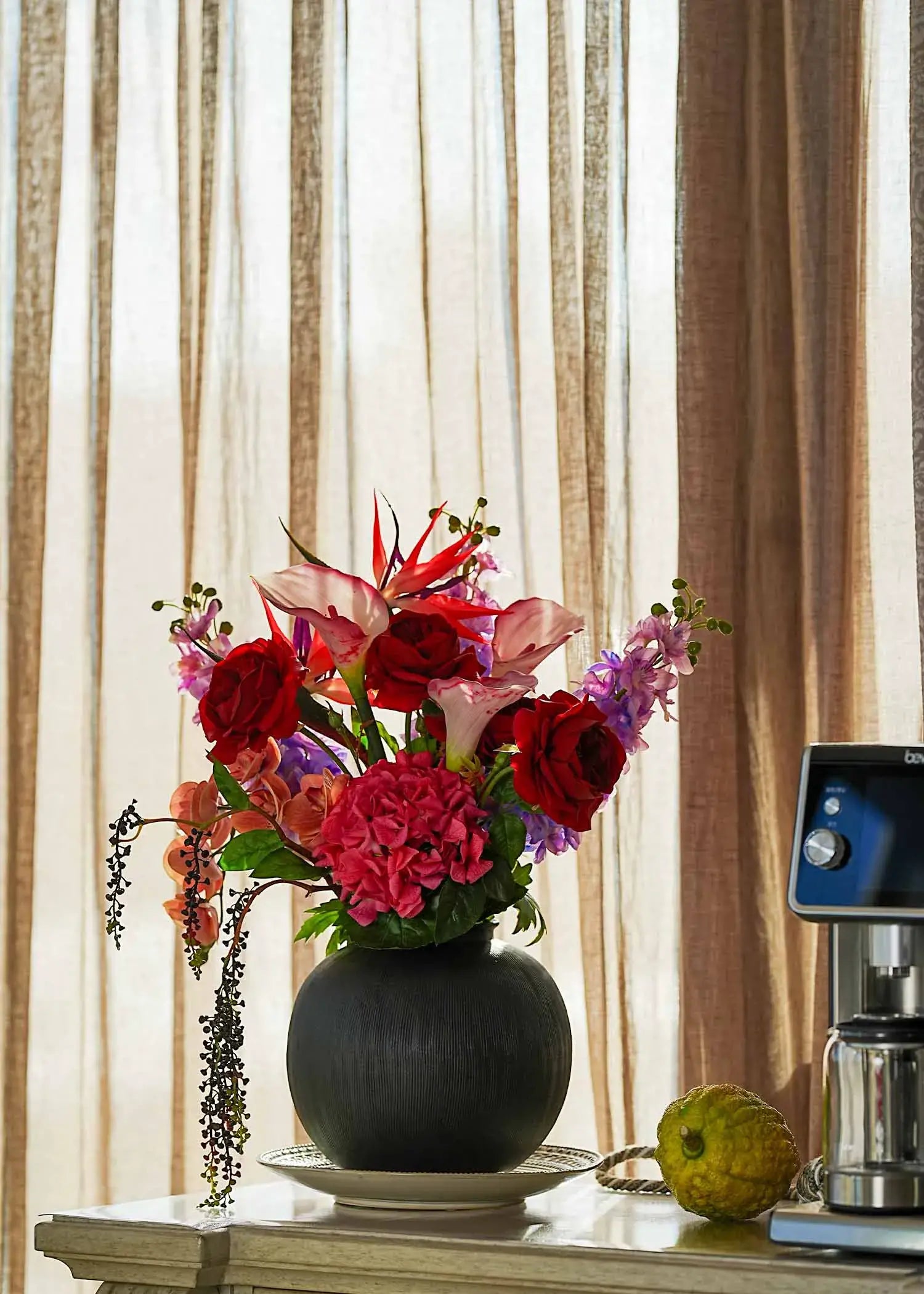 Crimson Paradise faux flower bouquet in a black vase on a light-toned sideboard beside a coffee machine under soft daylight.