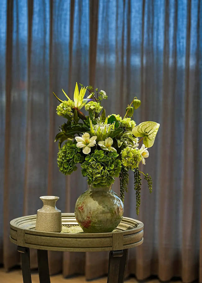 Ethereal Elegance artificial flower bouquet with green hydrangeas, lilies, and tropical leaves in a ceramic vase on a round table, elegant home décor accent.