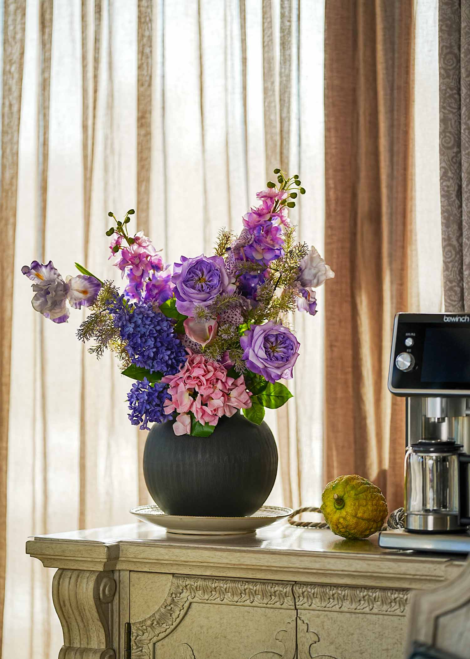 Lavender Whispers faux floral arrangement with purple tulips, irises, and roses in a dark ceramic vase on a cream cabinet beside a coffee machine, soft daylight setting.