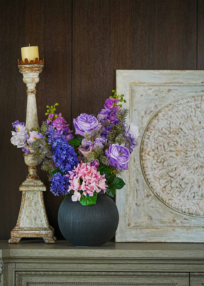 Lavender Whispers faux floral arrangement featuring purple roses, hydrangeas, and tulips in a matte black vase displayed on a vintage shelf beside a rustic candle stand.