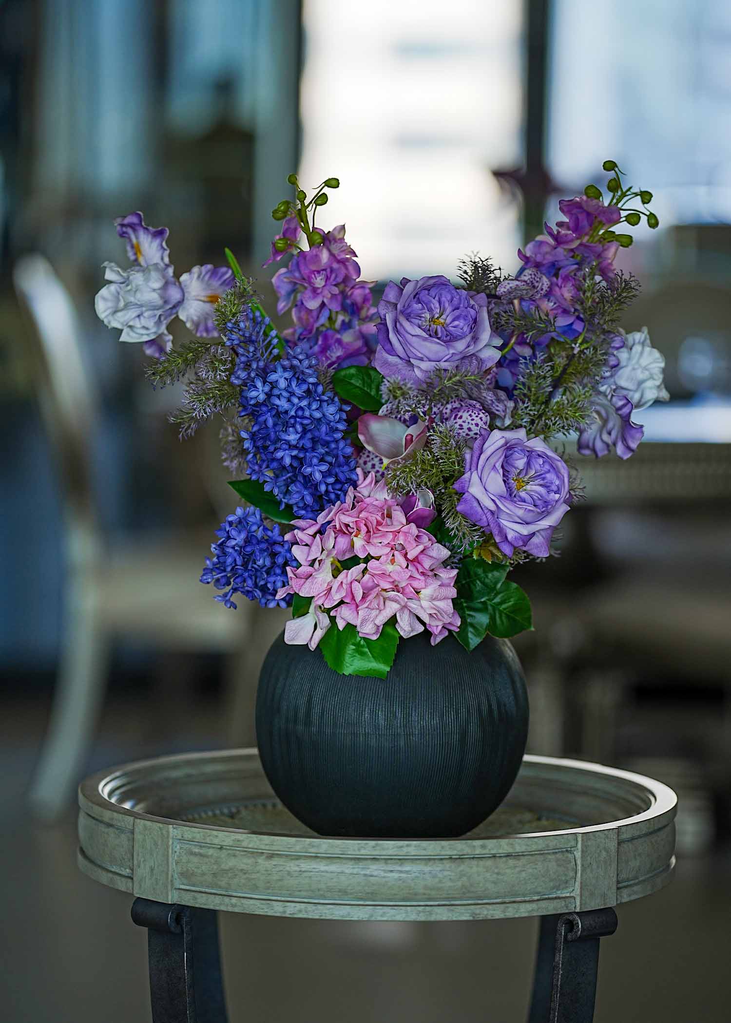 Lavender Whispers faux floral arrangement featuring purple roses, tulips, and hydrangeas in a dark ceramic vase displayed on a wooden round table in soft natural light.