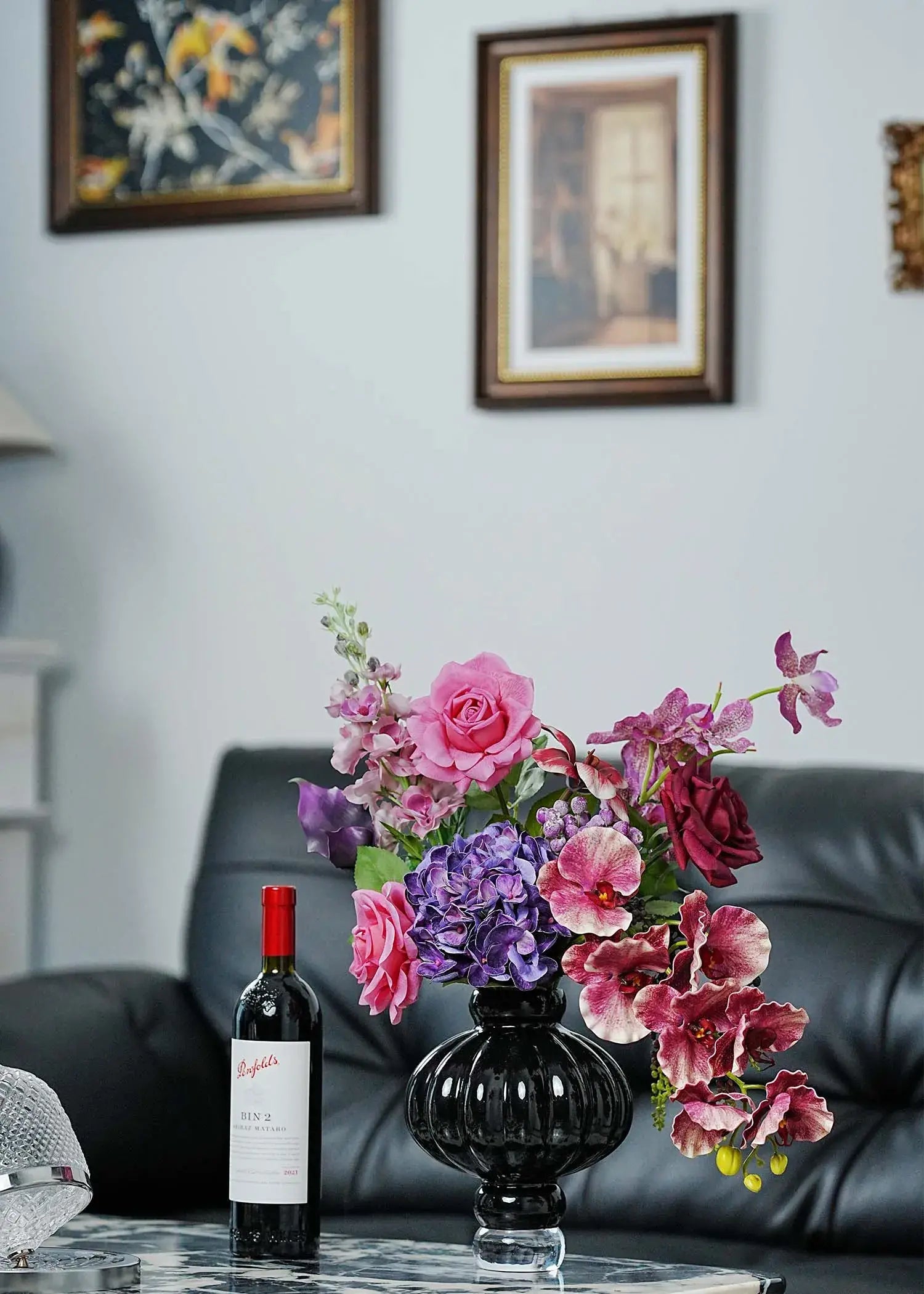 Luxury faux floral arrangement featuring pink roses, purple orchids, and burgundy calla lilies in a black vase, displayed on a marble table beside a bottle of red wine in a modern living room setting.
