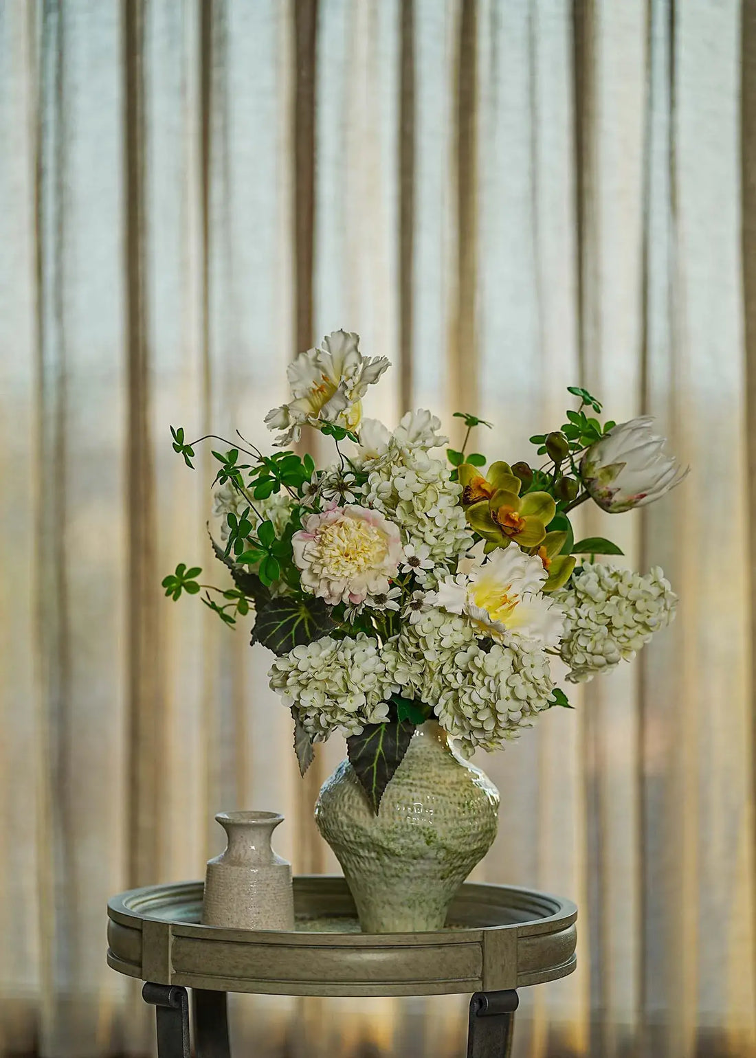 White hydrangea and peony faux floral arrangement in a textured green ceramic vase, displayed on a wooden table with soft natural light.