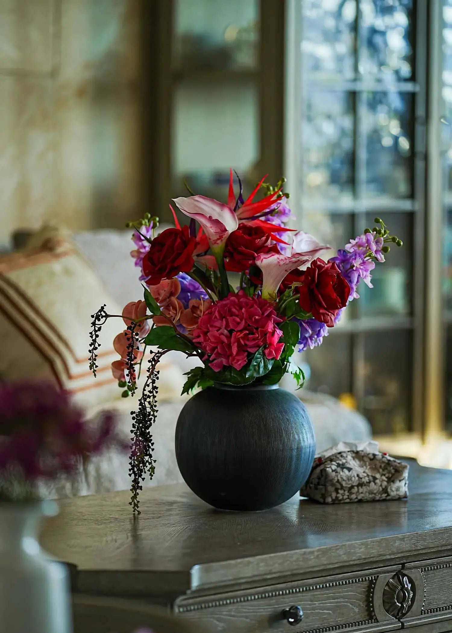 Crimson Paradise faux flower arrangement in a black vase displayed on a wooden coffee table in a cozy living room.