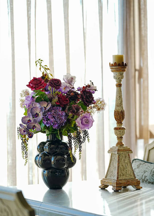 Deep purple and red artificial flower centerpiece with orchids and roses arranged in a glossy black vase.