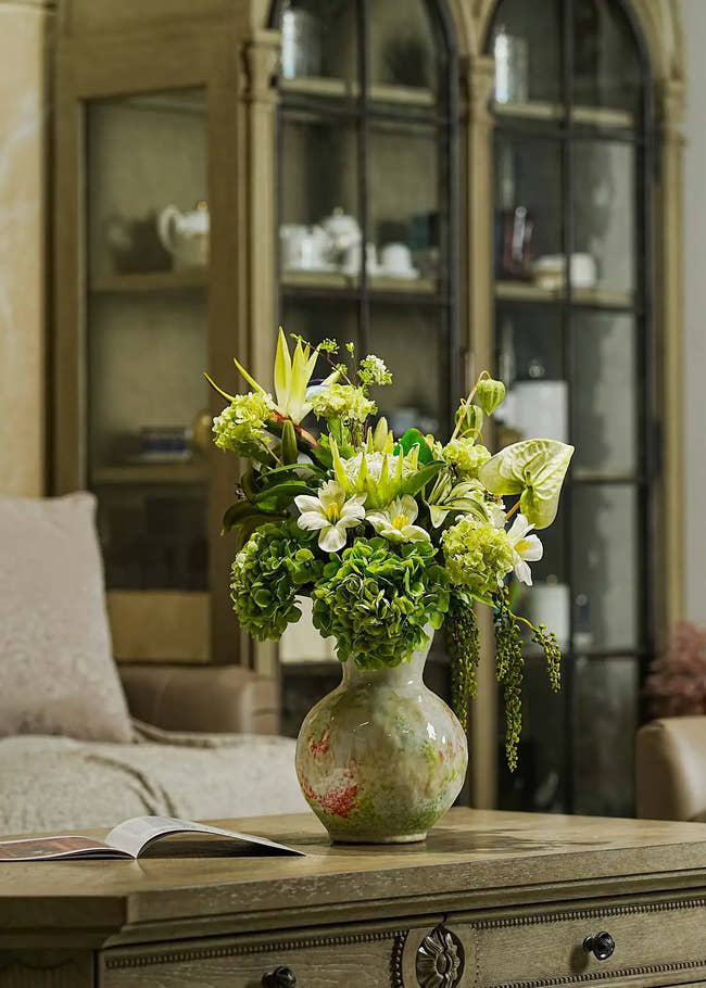 Ethereal Elegance artificial flower bouquet with green hydrangeas, lilies, and tropical leaves arranged in a ceramic vase on a wooden table in a cozy living room.