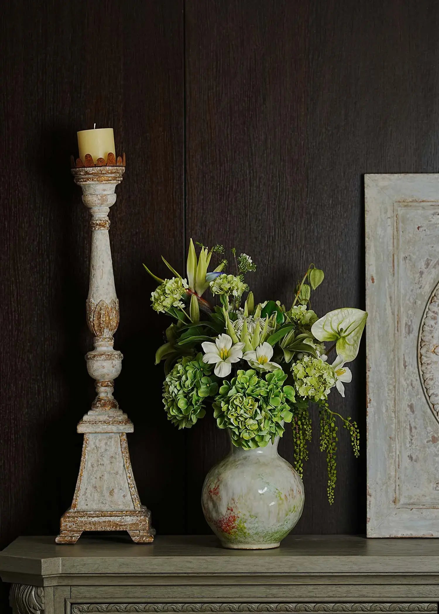 Ethereal Elegance artificial flower bouquet with green hydrangeas, white lilies, and tropical foliage in a ceramic vase displayed on a vintage shelf beside a candle stand.