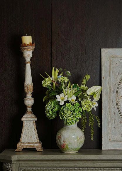 Ethereal Elegance artificial flower bouquet with green hydrangeas, white lilies, and tropical foliage in a ceramic vase displayed on a vintage shelf beside a candle stand.