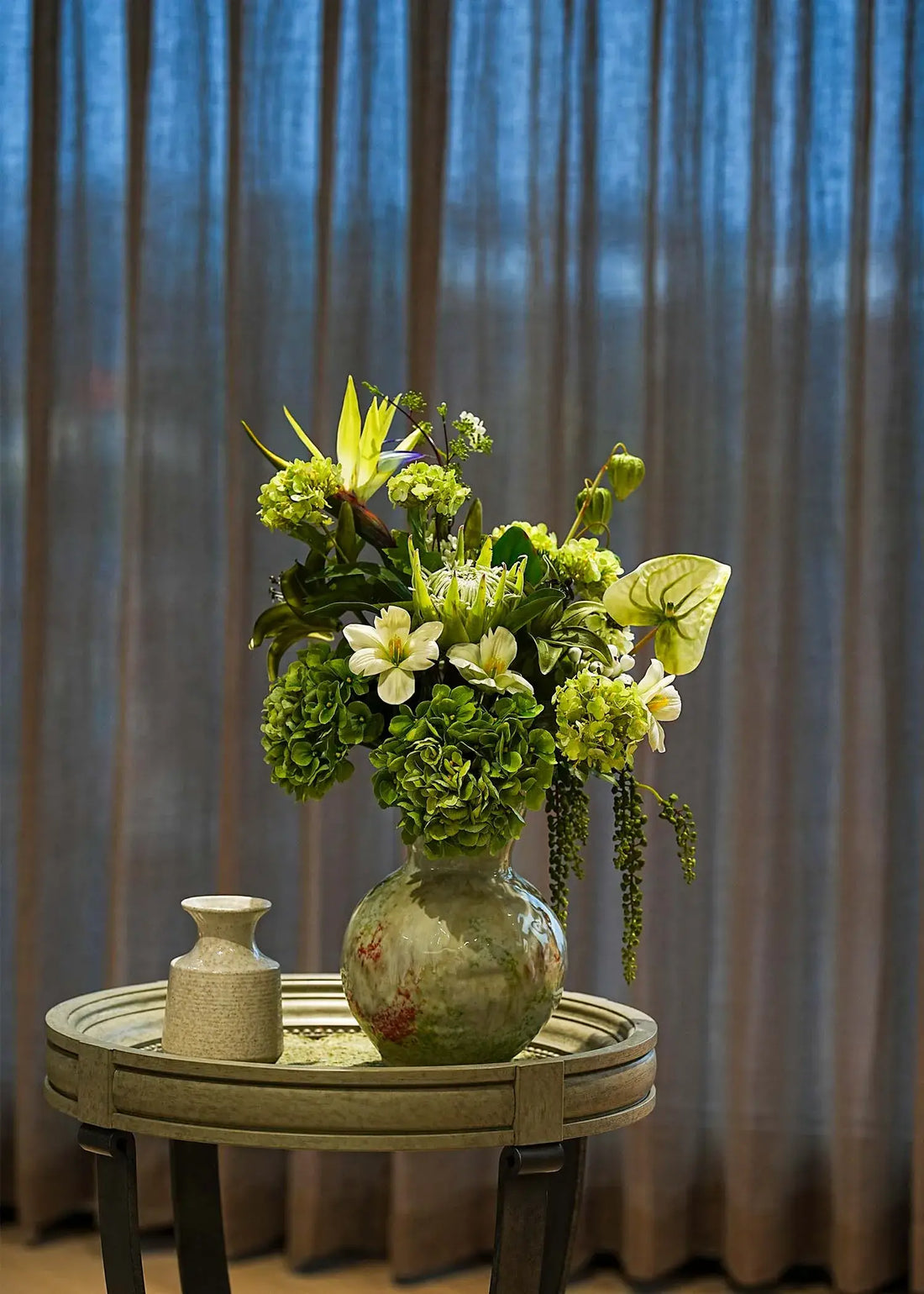 Ethereal Elegance artificial flower bouquet with green hydrangeas, lilies, and tropical leaves in a ceramic vase on a round table, elegant home décor accent.