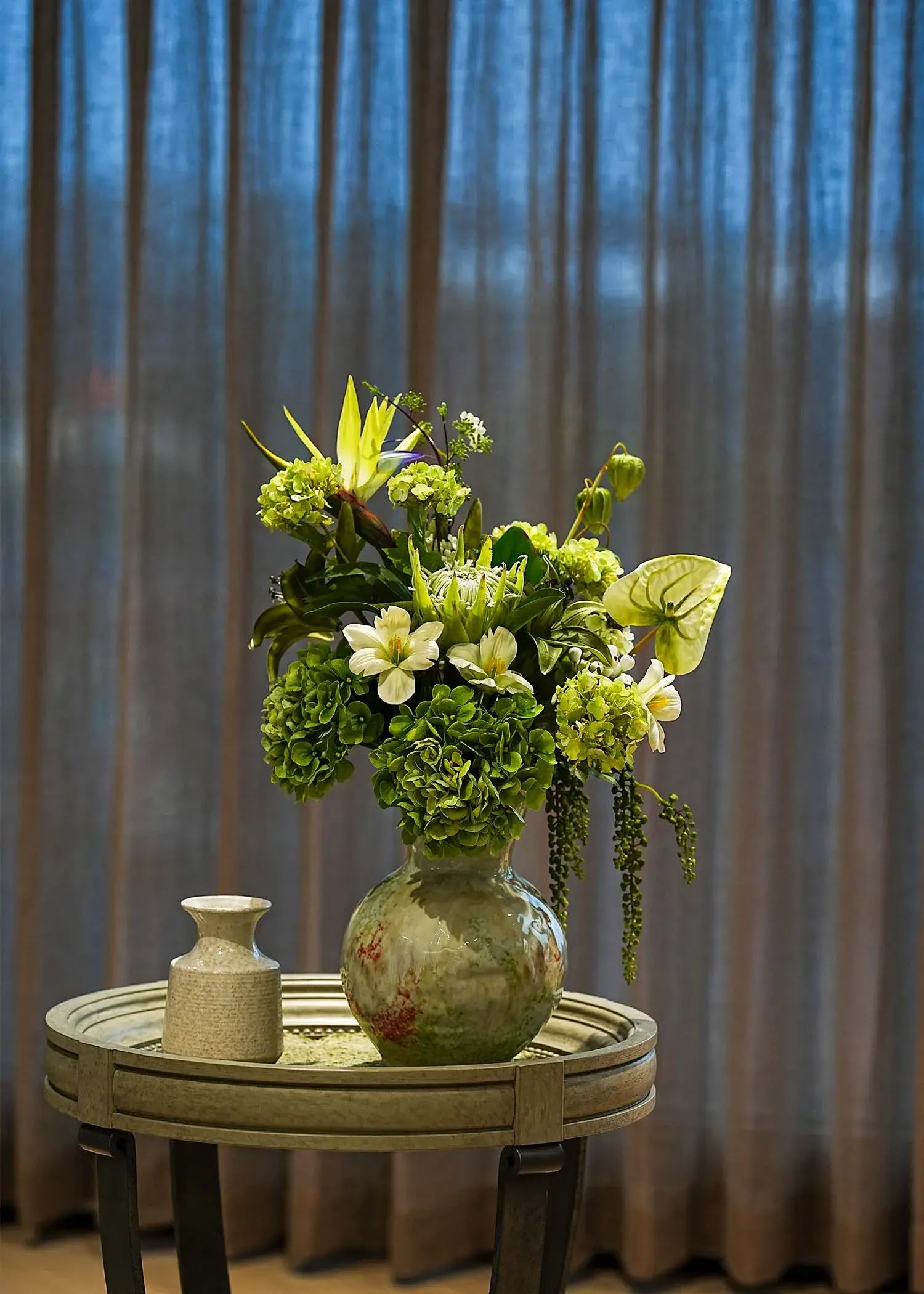 Ethereal Elegance artificial flower bouquet with green hydrangeas, lilies, and tropical leaves in a ceramic vase on a round table, elegant home décor accent.