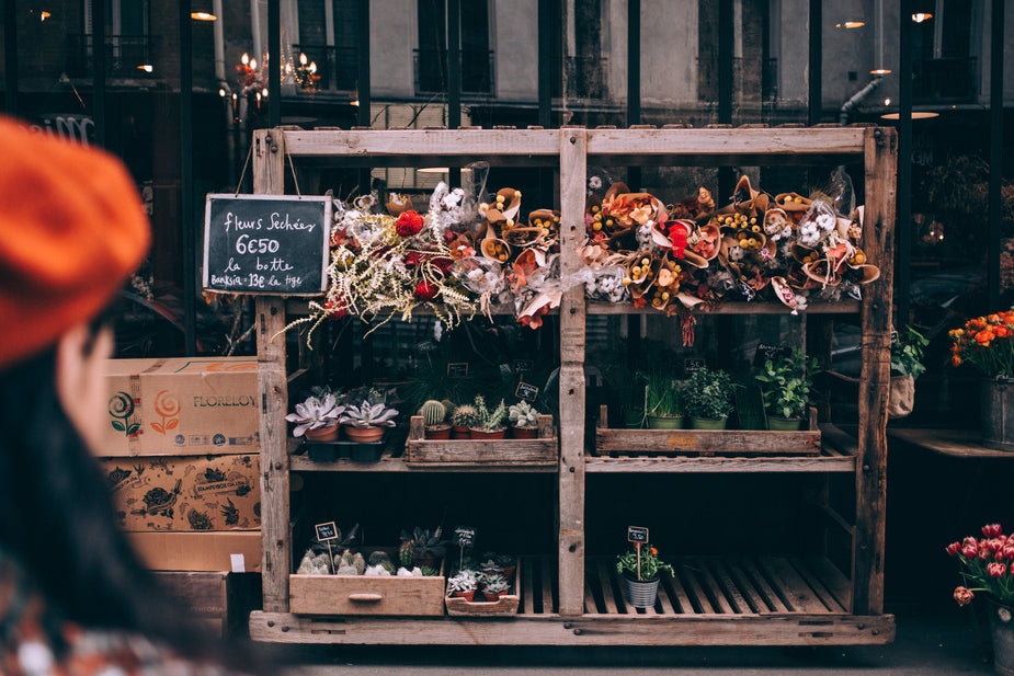 Rustic floral boutique storefront with wooden shelves, wrapped bouquets and potted succulents; chalkboard sign visible.