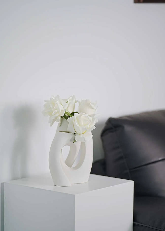 White artificial roses arrangement in abstract ceramic vase on white stand, minimalist living room decoration