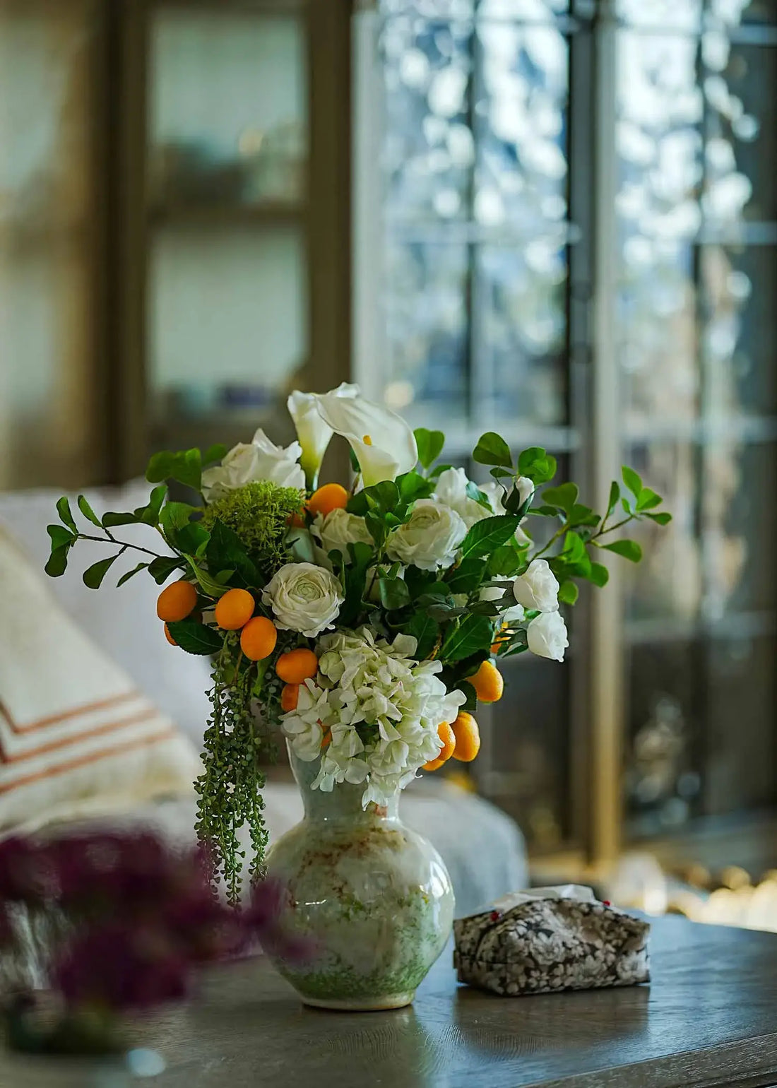 Lifelike faux white rose and citrus flower arrangement in a ceramic vase displayed on a wooden coffee table in a cozy living room.