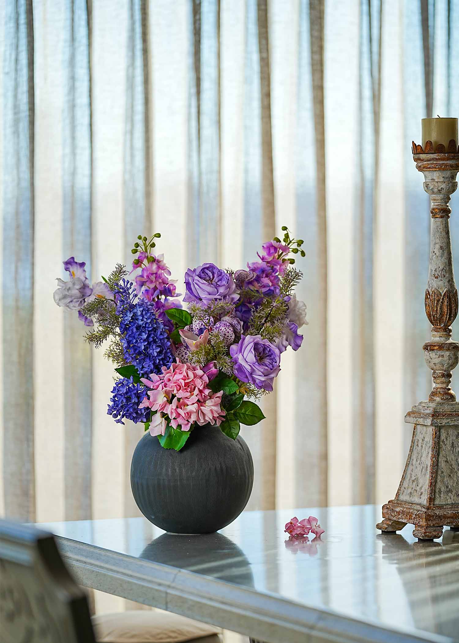 Lavender Whispers faux floral arrangement featuring purple roses, lilacs, and tulips in a dark ceramic vase placed on a dining table beside a rustic candle stand in daylight.