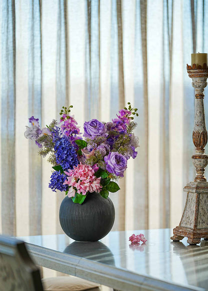 Lavender Whispers faux floral arrangement featuring purple roses, lilacs, and tulips in a dark ceramic vase placed on a dining table beside a rustic candle stand in daylight.