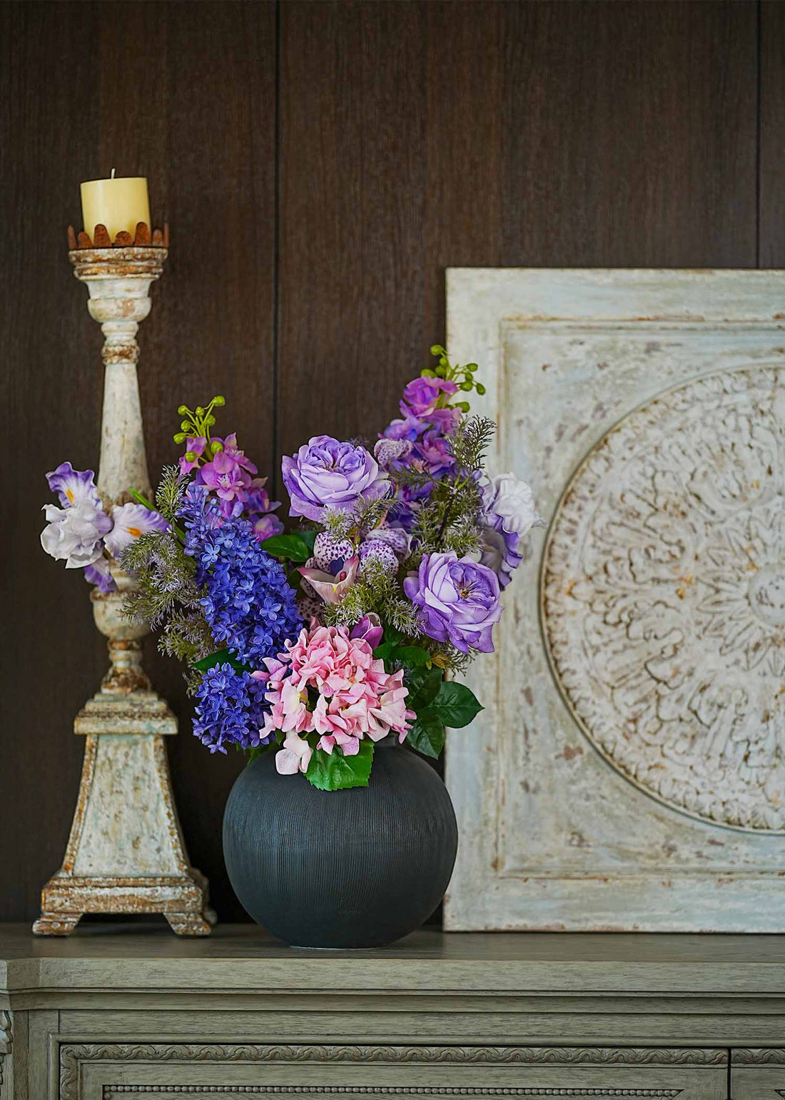 Lavender Whispers faux floral arrangement featuring purple roses, hydrangeas, and tulips in a matte black vase displayed on a vintage shelf beside a rustic candle stand.