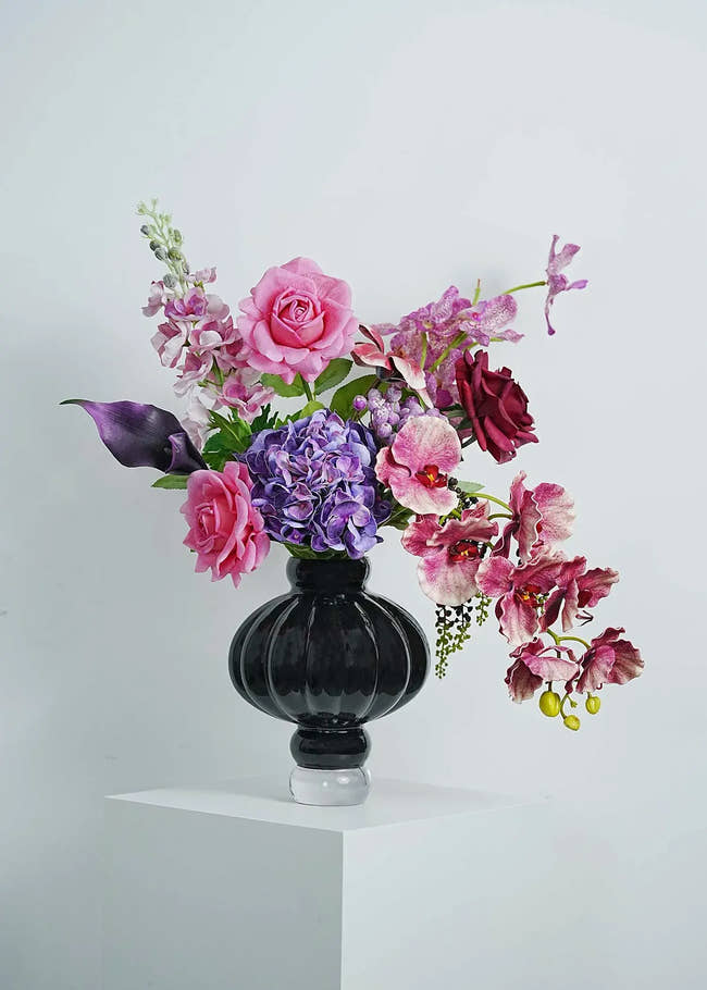 Luxury faux floral arrangement featuring pink roses, purple orchids, and dark calla lilies in an elegant black glass vase on a white pedestal.