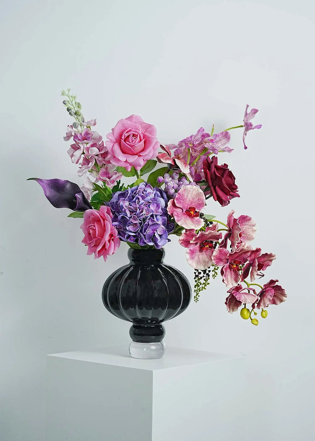 Luxury faux floral arrangement featuring pink roses, purple orchids, and dark calla lilies in an elegant black glass vase on a white pedestal.