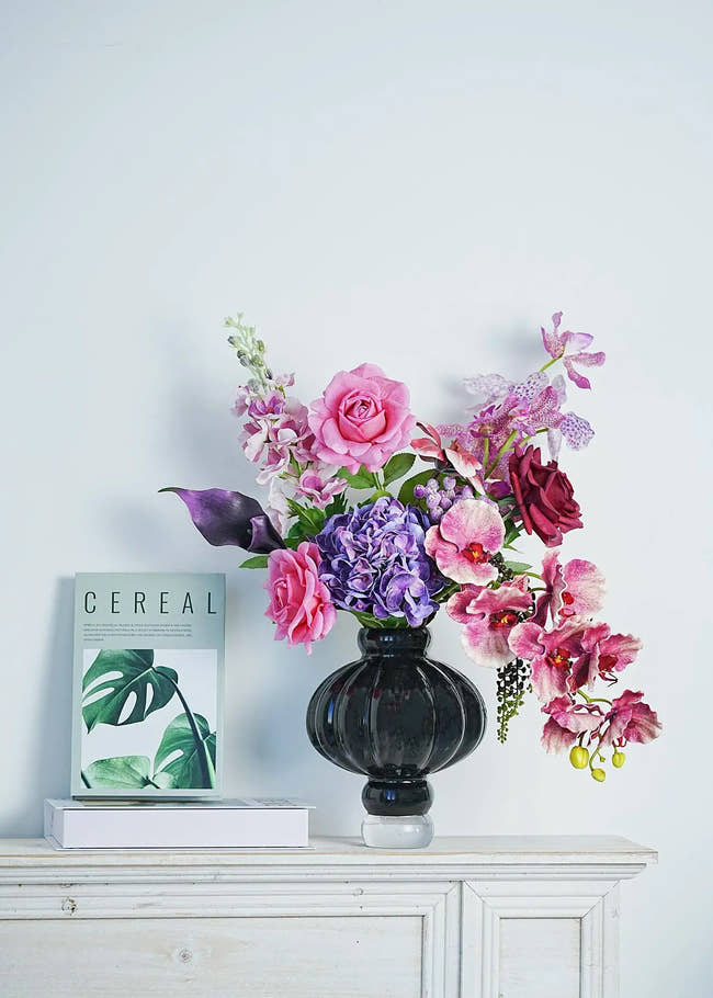 Luxury faux floral arrangement with pink roses, purple orchids, and burgundy calla lilies in a black vase, styled on a white console table beside a minimal magazine for home decor inspiration.