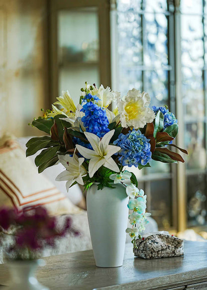 Luxury faux lily and hydrangea floral arrangement in a tall white vase placed on a cream dining cabinet beside a coffee machine, softly lit by natural sunlight.