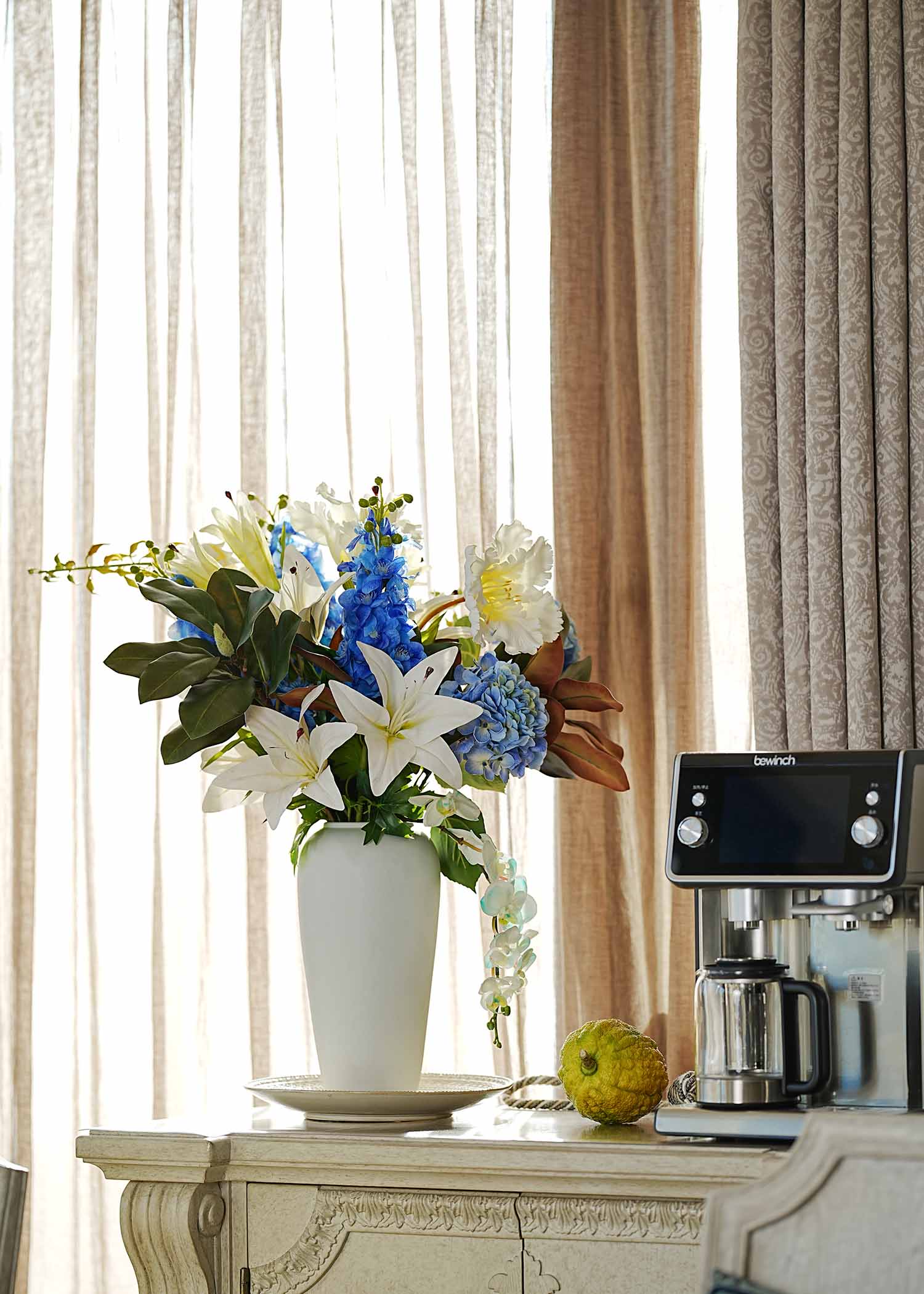 Luxury faux lily and hydrangea floral arrangement with blue and white blooms in a tall white vase placed on a wooden table in a cozy living room setting.