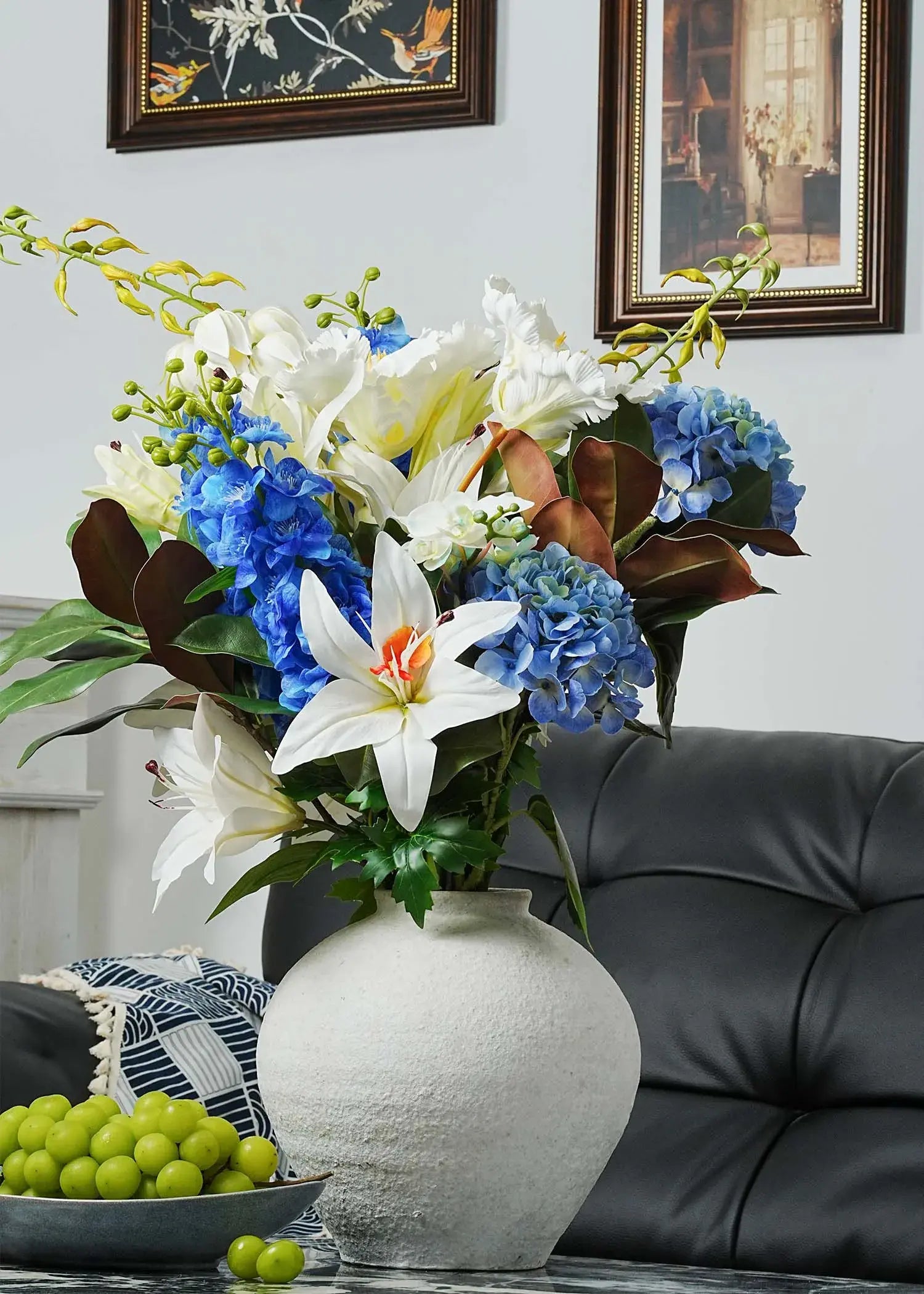 Luxury faux floral arrangement featuring white lilies, blue hydrangeas, and green foliage in a white ceramic vase, styled on a living room table with grapes and modern decor.