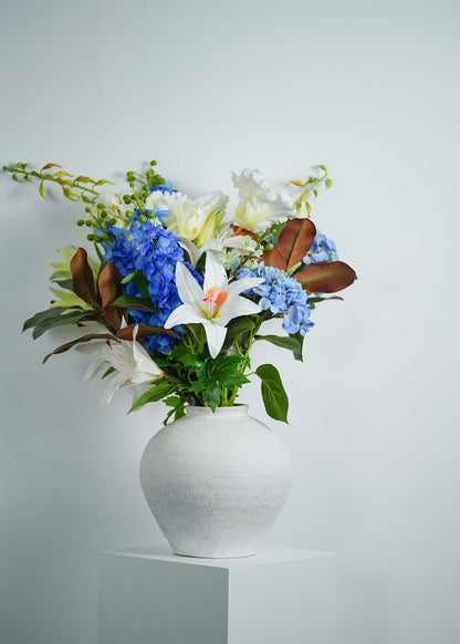 Luxury faux floral arrangement with white lilies, blue hydrangeas, and green leaves in a white ceramic vase on a minimalist pedestal.