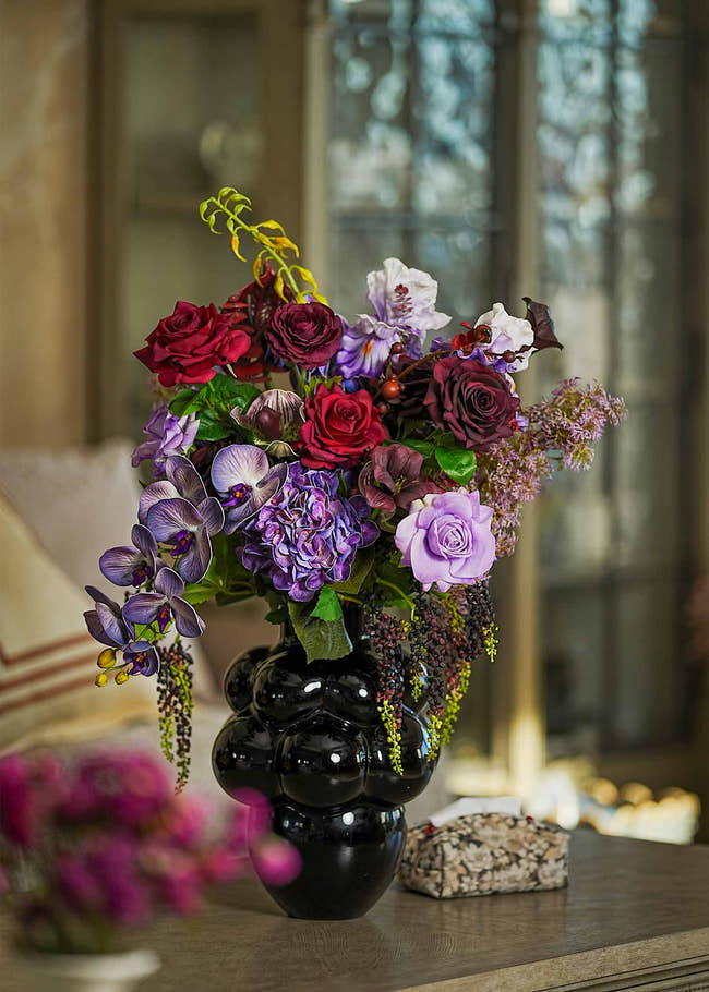 Royal Amethyst &amp; Burgundy faux floral arrangement featuring deep red roses, purple orchids, and hydrangeas in a sculptural black vase beside a candle holder.