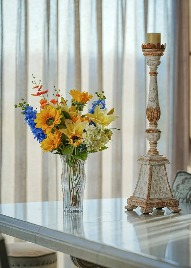 Sunlit Meadow faux sunflower and lily bouquet in a clear glass vase placed on a bright dining table beside a vintage candle holder.