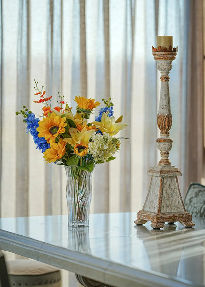 Sunlit Meadow faux sunflower and lily bouquet in a clear glass vase placed on a bright dining table beside a vintage candle holder.