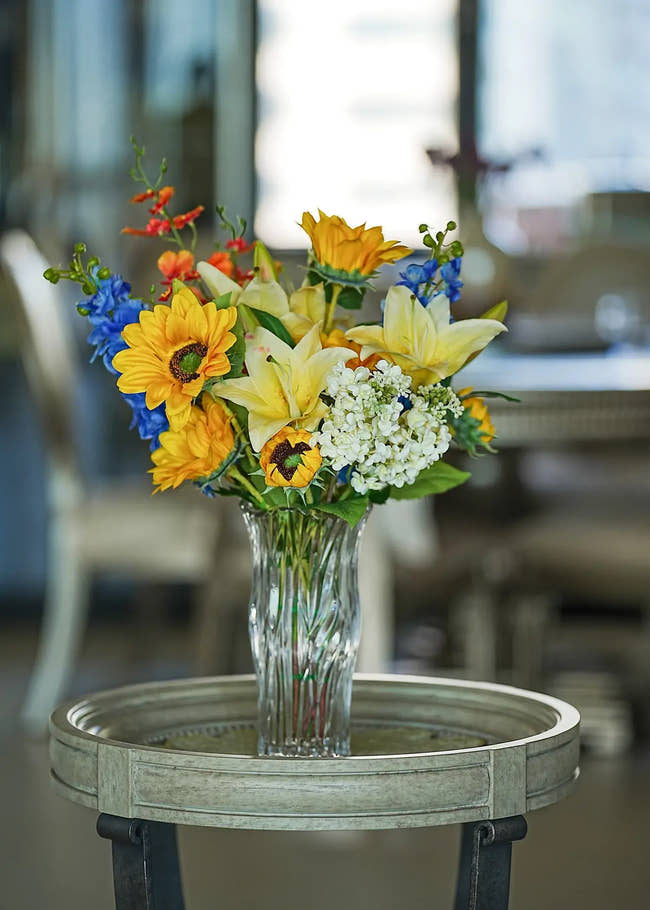 Sunlit Meadow faux sunflower and lily bouquet arranged in a clear glass vase on a wooden coffee table in a cozy living room setting.