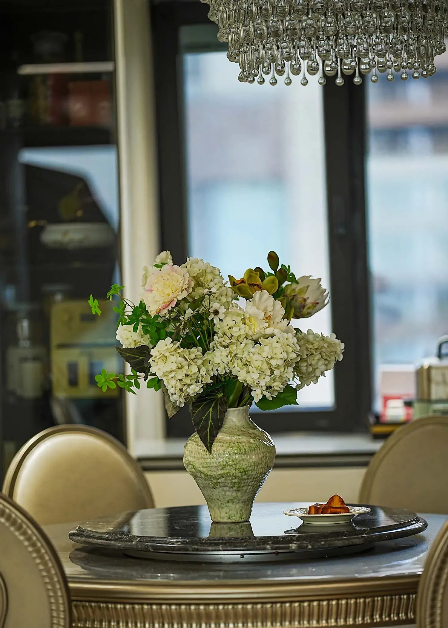 White hydrangea and peony faux floral arrangement in a ceramic vase displayed on a round dining table under a crystal chandelier, elegant home décor scene.