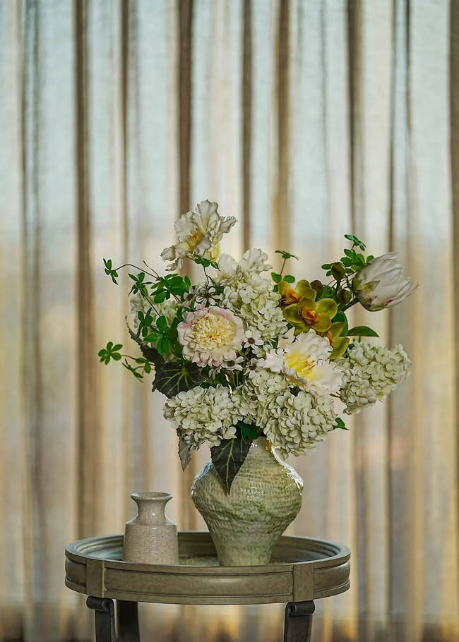 White hydrangea and peony faux floral arrangement in a textured green ceramic vase, displayed on a wooden table with soft natural light.